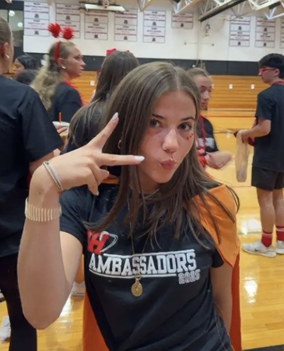 Tatum at a school event in an orange cape and an Ambassadors 2025 shirt, flashing a peace sign in the gym