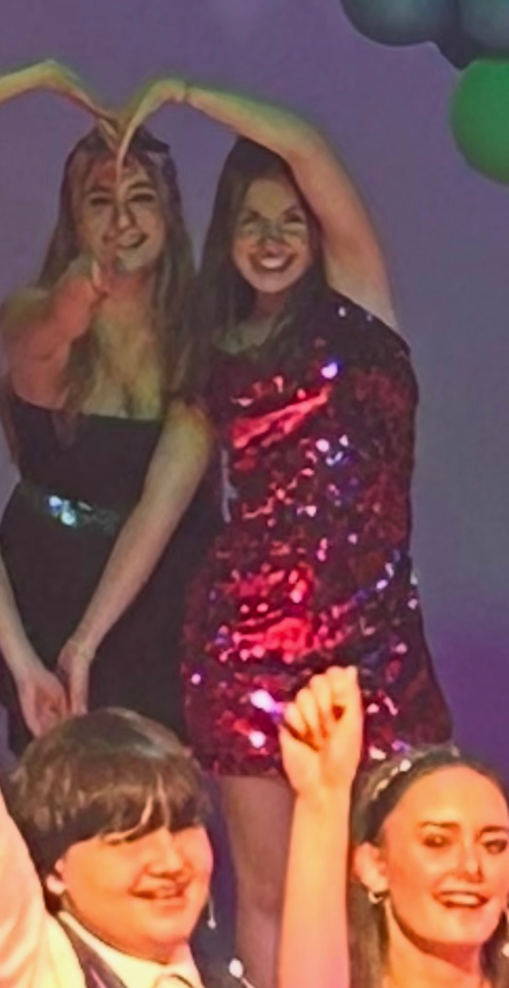 Tatum and a friend making a heart with their hands overhead at a party, balloons behind them