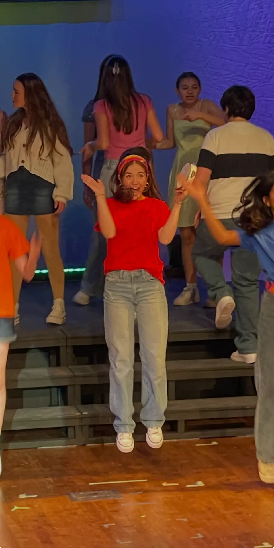 Tatum mid-jump in a red t-shirt on a stage riser, surrounded by the cast
