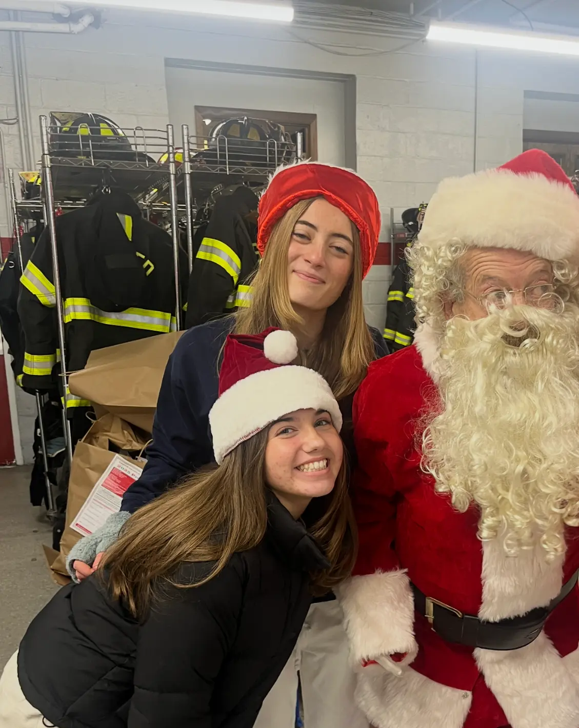 Tatum and a classmate with Santa Claus inside the firehouse, surrounded by gift bags and turnout gear